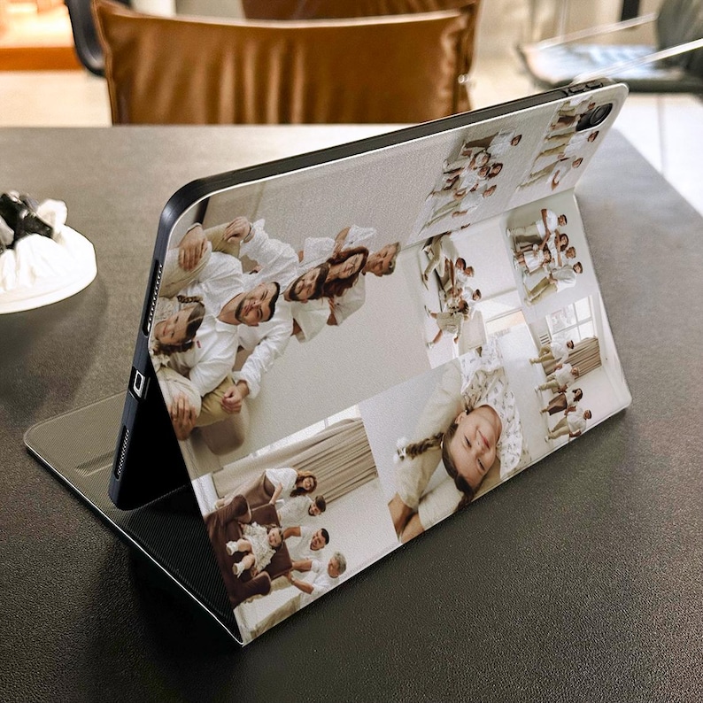 Puede incluir: Una funda negra para tableta con un dise&ntilde;o de collage de fotos. El collage presenta m&uacute;ltiples fotos familiares en una variedad de tama&ntilde;os y orientaciones. La funda est&aacute; abierta y apoyada, mostrando las im&aacute;genes. El fondo es una superficie oscura.