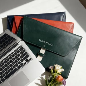 May include: Leather document holders in navy, orange, and green. The green holder has the text "YOUR NAME". A laptop and a small bouquet of flowers are also in the image.