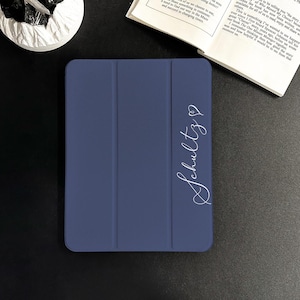Puede incluir: Una funda para tableta azul marino con un diseño plegable. La funda presenta el nombre "Schultz" en una fuente blanca con un símbolo de corazón. La funda está cerrada y parece estar hecha de un material liso.