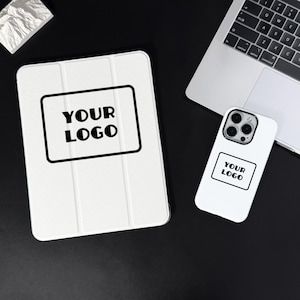 Puede incluir: Funda blanca para tableta y funda para teléfono con el texto "YOUR LOGO" en un rectángulo negro. La funda de la tableta está plegada y tiene una superficie texturizada. Un portátil está en el fondo.
