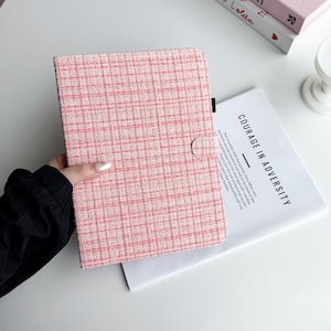May include: Pink and white plaid tablet case with a button closure. The case is made of a textured fabric. A magazine with the words "COURAGE IN ADVERSITY" is visible in the background.