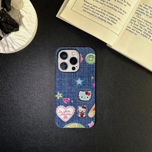 May include: A blue denim iPhone case with a Hello Kitty design. The case features a variety of colorful patches, including stars, hearts, and a Hello Kitty face. The text "In your dreams" is printed on a pink heart patch.