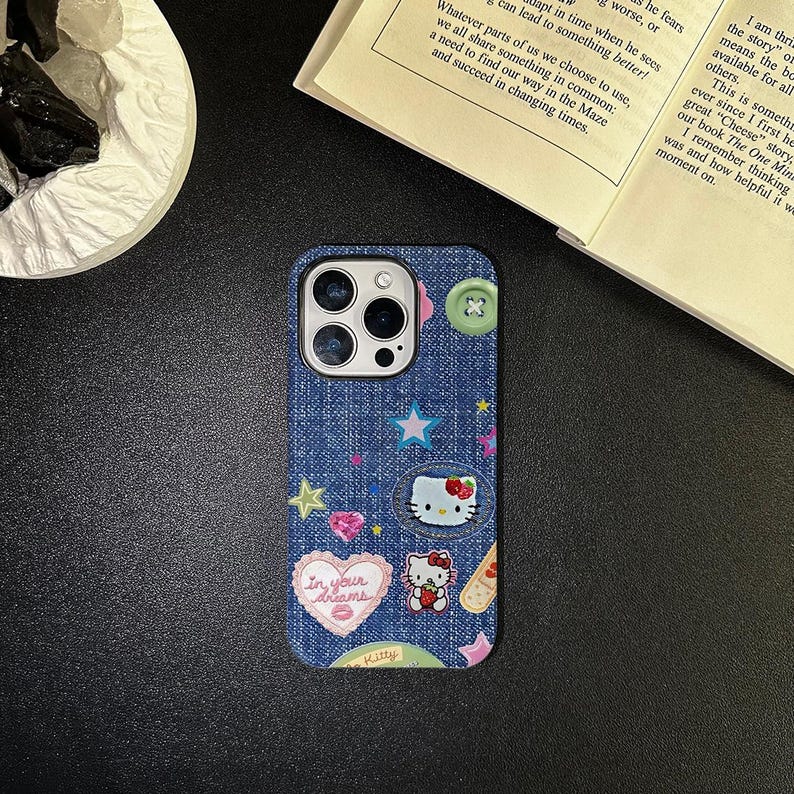May include: A blue denim iPhone case with a Hello Kitty design. The case features a variety of colorful patches, including stars, hearts, and a Hello Kitty face. The text "In your dreams" is printed on a pink heart patch.