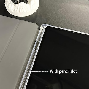 Puede incluir: Una funda protectora transparente para tablet con ranura para l&aacute;piz. La funda est&aacute; parcialmente abierta, mostrando la pantalla de la tablet. Tiene una cubierta gris y un marco transparente. El texto "With pencil slot" es visible.
