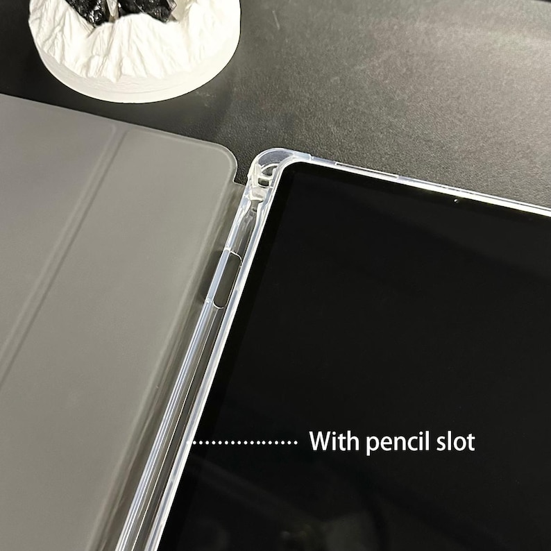 Puede incluir: Una funda protectora transparente para tablet con ranura para l&aacute;piz. La funda est&aacute; parcialmente abierta, mostrando la pantalla de la tablet. Tiene una cubierta gris y un marco transparente. El texto "With pencil slot" es visible.