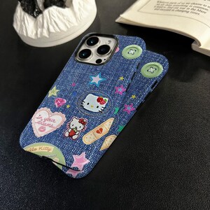 May include: A blue denim iPhone case with a Hello Kitty design. The case features a variety of colorful cartoon characters, stars, and hearts. The text "In your dreams" is printed on a pink heart.