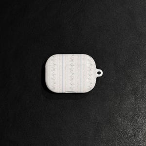 May include: White AirPod case with a floral pattern in pink and blue. The case has a vertical striped design and a small loop for attaching to a keychain. The case is on a black surface.