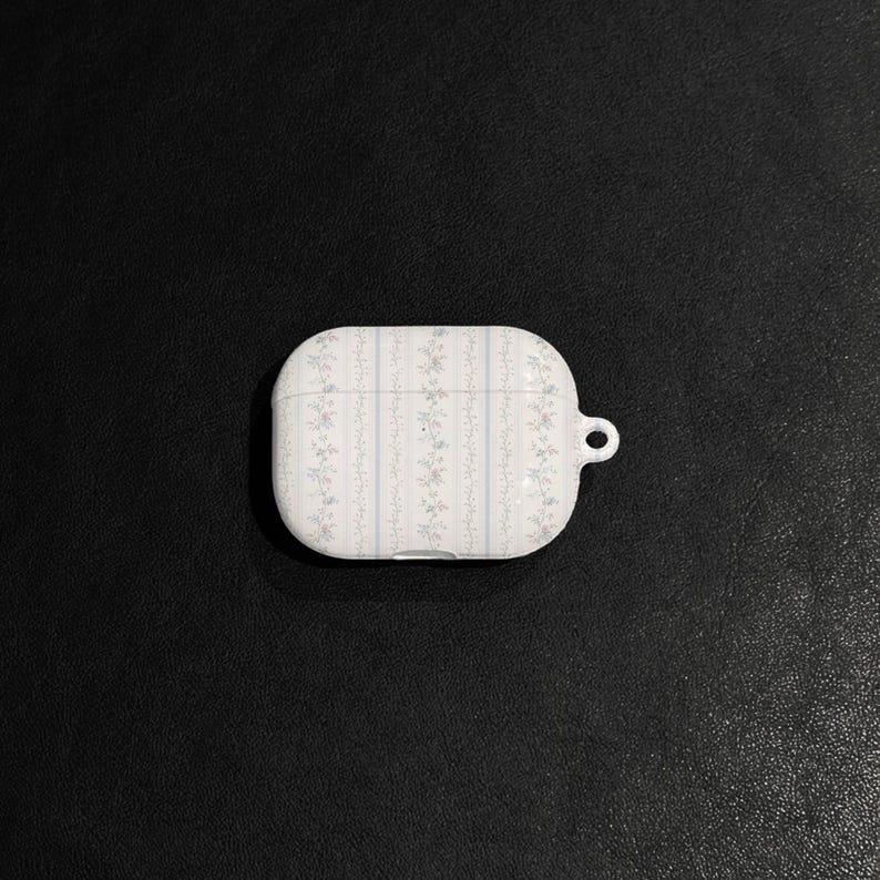 May include: White AirPod case with a floral pattern in pink and blue. The case has a vertical striped design and a small loop for attaching to a keychain. The case is on a black surface.