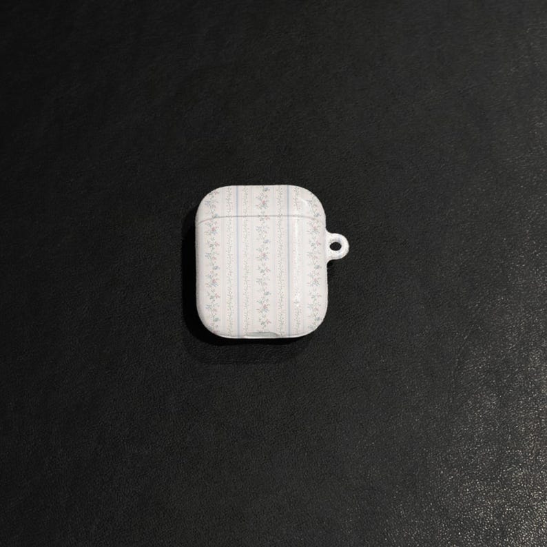 May include: White AirPod case with a floral pattern in light blue and gray. The case has a small loop for attaching to a keychain. The case is rectangular with rounded edges.