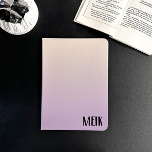 May include: A small, rectangular notebook with a colour gradient, shifting from light beige to lavender. The word "MEIK" is printed in black at the bottom right corner. The notebook is on a black surface, with a book and decorative glass pieces in the background.