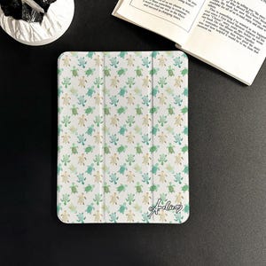 Può includere: Custodia bianca per tablet con un motivo di tartarughe marine ad acquerello nei toni del verde e del blu. La custodia ha una copertura pieghevole e una firma in corsivo. Un libro aperto è visibile sullo sfondo.