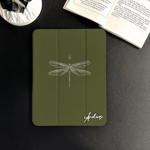 Puede incluir: Funda de tableta verde oliva con una ilustración de libélula blanca y el nombre "Amelie" en cursiva. La funda está cerrada, con una solapa magnética. El diseño de la libélula está centrado en la parte delantera.