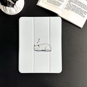 Può includere: Custodia bianca per tablet con un'illustrazione minimalista di un gatto che dorme. Il gatto è accompagnato dalle lettere "Z" sopra. La custodia ha un design a tre ante.