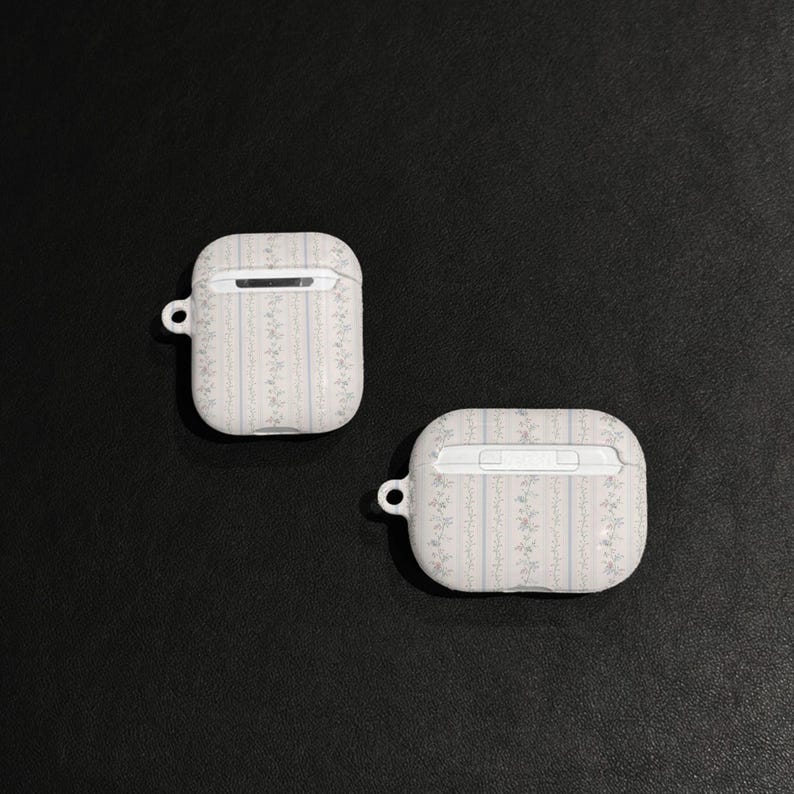 May include: Two white AirPod cases with a floral pattern and a small loop for a keychain. The cases have a rectangular shape with rounded edges and a small opening at the top.