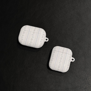 May include: Two white AirPod cases with a floral pattern in light blue and gray. The cases are rectangular with rounded edges and a small loop for attaching a lanyard. The background is a dark gray surface.