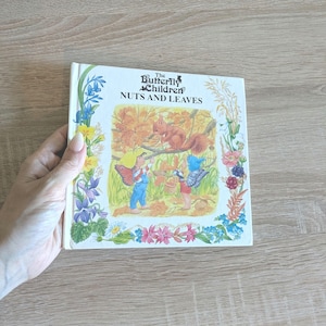 Puede incluir: Un libro infantil vintage titulado "The Butterfly Children Nuts and Leaves". La portada presenta una ilustración de una ardilla, niños y hojas de otoño. El libro está sostenido por una mano y está sobre una superficie de madera clara.