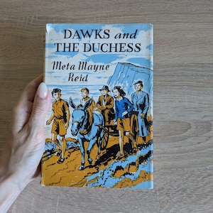 May include: Vintage book titled "DAWKS and THE DUCHESS" by Meta Mayne Reid. The cover features an illustration of a group of people with a donkey cart, set against a blue sky and coastal backdrop. The book is held by a person.