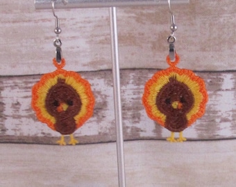 May include: A pair of crocheted turkey earrings with orange, brown, and yellow accents hanging on a silver earring stand.