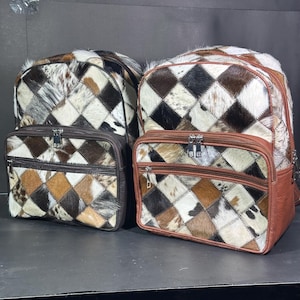 Pode incluir: Duas mochilas de couro marrom com um padrão de diamante feito de diferentes pedaços de couro de vaca de cores diferentes. As mochilas têm dois compartimentos com zíper e um bolso menor com zíper na frente.