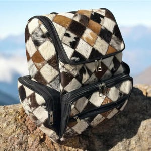 May include: A patchwork backpack with a geometric pattern of brown, black, and white squares. The bag has multiple zippered compartments and black leather trim. The backpack is sitting on a rock with a mountain backdrop.