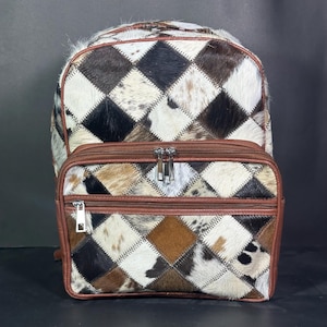 Puede incluir: Un juego de mochila y bolsa a juego. Las bolsas están hechas de un diseño de patchwork con cuero de vaca marrón, negro y blanco. La mochila tiene una parte superior redondeada y correas ajustables. La bolsa tiene un cierre de cremallera y un ribete de cuero marrón.