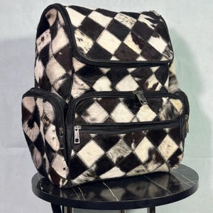 May include: A black and white cowhide backpack with a diamond pattern. The backpack has multiple zippered compartments and black leather trim. The bag is sitting on a black surface.