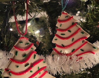 Pode incluir: Duas decorações de árvore de Natal. Cada árvore é branca com listras de glacê vermelho e granulado verde. Os enfeites penduram-se em fios vermelhos e verdes, expostos numa árvore de Natal com enfeites brancos.
