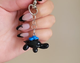 May include: A black turtle keychain with a blue hat charm. The keychain is attached to a silver chain and a silver lobster clasp.