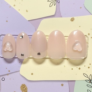 May include: A set of five oval-shaped, light pink press-on nails. Two nails feature small, clear heart-shaped embellishments, and two have silver rhinestones. The nails are displayed on a clear plastic holder against a pastel background with decorative elements.