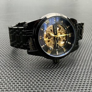 May include: A black and gold automatic wristwatch with a skeleton dial. The watch has a black metal band and a clear face with Roman numeral hour markers.