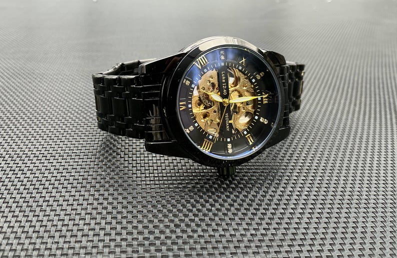 May include: A black and gold automatic wristwatch with a skeleton dial. The watch has a black metal band and a clear face with Roman numeral hour markers.
