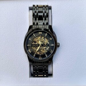 May include: A black automatic wristwatch with a gold-tone face and Roman numeral hour markers. The watch has a skeletonized design, revealing the inner workings of the movement. The watch face also features the text "Luke 12-40" and "YEDOVANY".