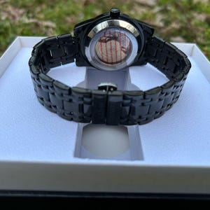 May include: A black wristwatch with a black metal band and a round face. The watch is in a white box.