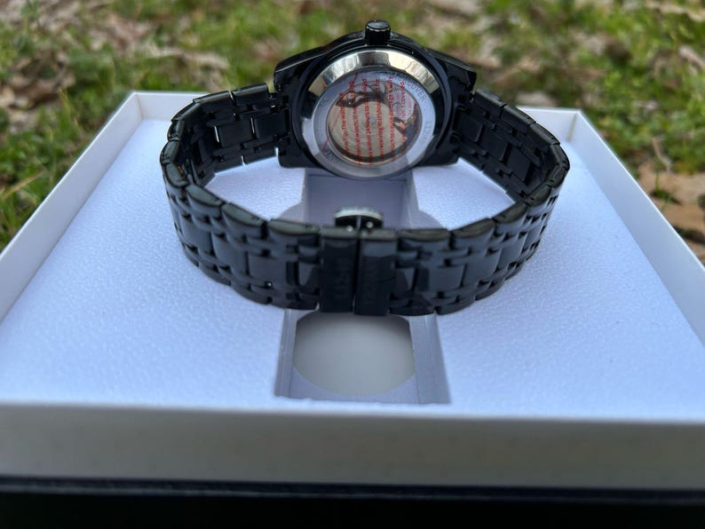 May include: A black wristwatch with a black metal band and a round face. The watch is in a white box.