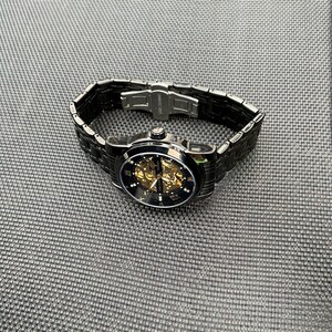 May include: A black automatic wristwatch with a gold skeleton movement and black Roman numeral hour markers. The watch has a black metal band with a clasp closure.