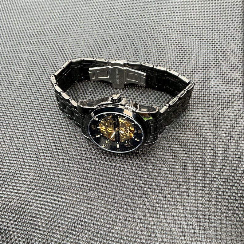 May include: A black automatic wristwatch with a gold skeleton movement and black Roman numeral hour markers. The watch has a black metal band with a clasp closure.