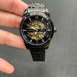 May include: Black automatic wristwatch with a gold skeleton movement and a black metal band. The watch face has Roman numeral hour markers and a small seconds dial. The watch face also has the text "Luke 12:40" and "Yeovany".