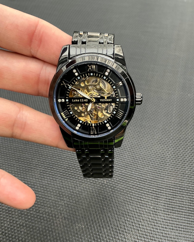May include: Black automatic wristwatch with a gold skeleton movement and a black metal band. The watch face has Roman numeral hour markers and a small seconds dial. The watch face also has the text "Luke 12:40" and "Yeovany".