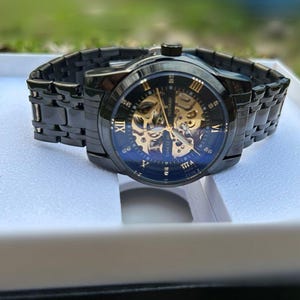 May include: A black and gold automatic wristwatch with a skeletonized dial and Roman numeral hour markers. The watch has a black metal band and is displayed in a white box.