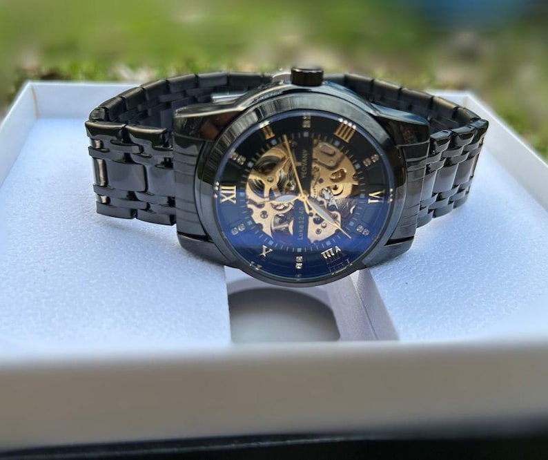 May include: A black and gold automatic wristwatch with a skeletonized dial and Roman numeral hour markers. The watch has a black metal band and is displayed in a white box.
