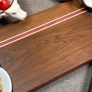 May include: A rectangular walnut wood cutting board with a purple and white wood inlay. The board has a smooth, polished surface.