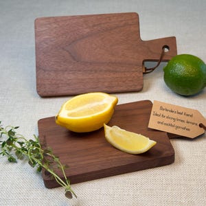 May include: Two small walnut wood cutting boards with a lemon and a lime wedge on one of the boards. The boards are ideal for slicing limes, lemons, and cocktail garnishes. The text on the tag reads "Bartender's best friend. Ideal for slicing limes, lemons, and cocktail garnishes."