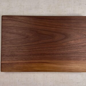 May include: A rectangular wooden cutting board with a rich, dark brown color and visible wood grain. The board has a smooth surface and a natural, warm tone, suitable for kitchen use.