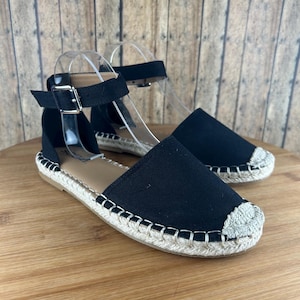 May include: Black canvas espadrille sandals with an ankle strap and buckle closure. The sandals have a white rope sole.