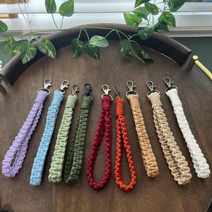 Puede incluir: Surtido de 10 llaveros de crochet de colores vivos con anillos metálicos. Los llaveros se muestran en una bandeja de madera. Los colores incluyen lavanda, azul claro, verde, verde oscuro, rojo, naranja, beige y blanco.