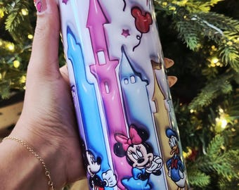 Op de afbeelding: Een witte beker met een kleurrijk kasteelontwerp met Mickey Mouse, Minnie Mouse en Donald Duck. De beker heeft een zilveren deksel en rietje en is versierd met sterren, ballonnen en vuurwerk. De beker wordt in een hand gehouden.