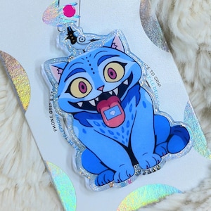 May include: A blue PopSockets phone grip featuring a cartoon cat with a pink tongue and large yellow eyes. The grip is attached to a white backing with the PopSockets logo and holographic accents. The background is a fluffy, white surface.