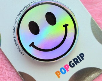 Op de afbeelding: Een witte PopSockets PopGrip met een holografisch smiley-gezichtsontwerp. De verpakking bevat de tekst "Phone Grip & Stand with Swappable Top - Try Me!  I'll  Stick  To  You!"