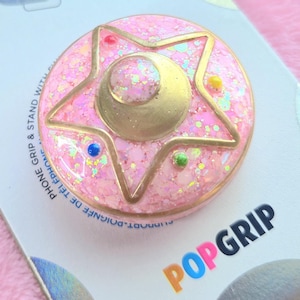 Op de afbeelding: Een roze en gouden telefoon grip met een glitter afwerking en een ster en halve maan ontwerp. De grip is gelabeld "POPGRIP" en "POPSOCKETS".