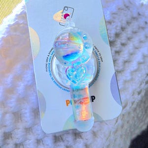 Op de afbeelding: Een transparante, iriserende telefoonhouder met een hart- en maanontwerp. De houder is verpakt op een witte kaart met de tekst "POPSOCKETS" en "TWICE TO GRIP" erop gedrukt.